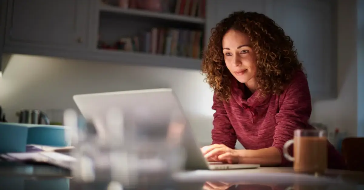 Blog 1 A person with curly hair leans over a laptop, surrounded by a mug and papers, illuminated by soft kitchen lighting.