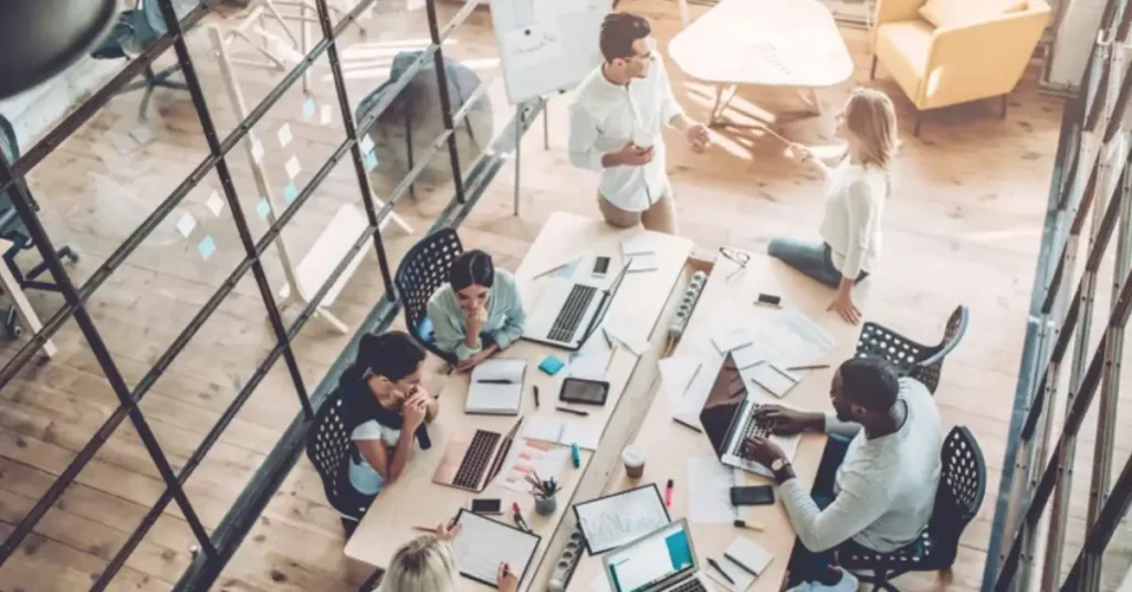A collaborative workspace where diverse individuals engage in teamwork, surrounded by laptops, documents, and creative materials on a wooden table.