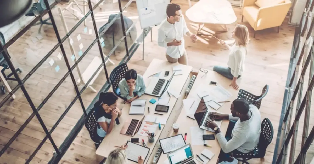 A collaborative workspace where diverse individuals engage in teamwork, surrounded by laptops, documents, and creative materials on a wooden table.